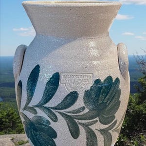 May include: Off-white stoneware vase with two handles and a flared rim. The vase features a teal floral design and the text "Salmon Falls Stoneware 1999". The vase is sitting on a rock with a scenic background.
