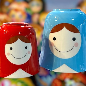 May include: Two ceramic cups with a smiling face design. The cups are shaped like a traditional Russian nesting doll. One cup is red and the other is blue.