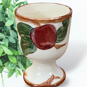 May include: A white ceramic egg cup with a brown rim and a red apple design. The apple has green leaves and a brown stem.
