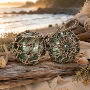 May include: Two clear glass fishing floats encased in a beige rope netting. The floats are connected by a rope handle. The background includes a beach scene with seashells and driftwood, evoking a coastal theme.
