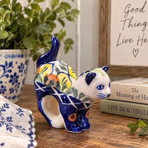 May include: A ceramic cat-shaped napkin ring featuring a glossy white base with hand-painted blue, yellow, and orange floral and lattice patterns. The cat is depicted in a standing position with its tail curved upward, showcasing intricate details.