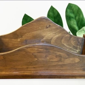 May include: A wooden desk organizer with a natural finish. The organizer has a sloped back and a curved front, with two compartments for storing mail or office supplies. Green leaves are visible in the background.