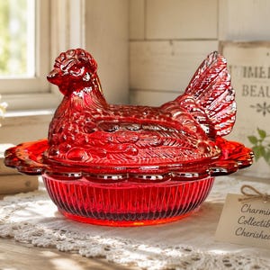 May include: A vibrant red glass hen on a nest dish featuring a detailed molded chicken lid with textured feather patterns and a scalloped edge base. The translucent ruby-colored glass captures light, showcasing the intricate relief design of the bird.