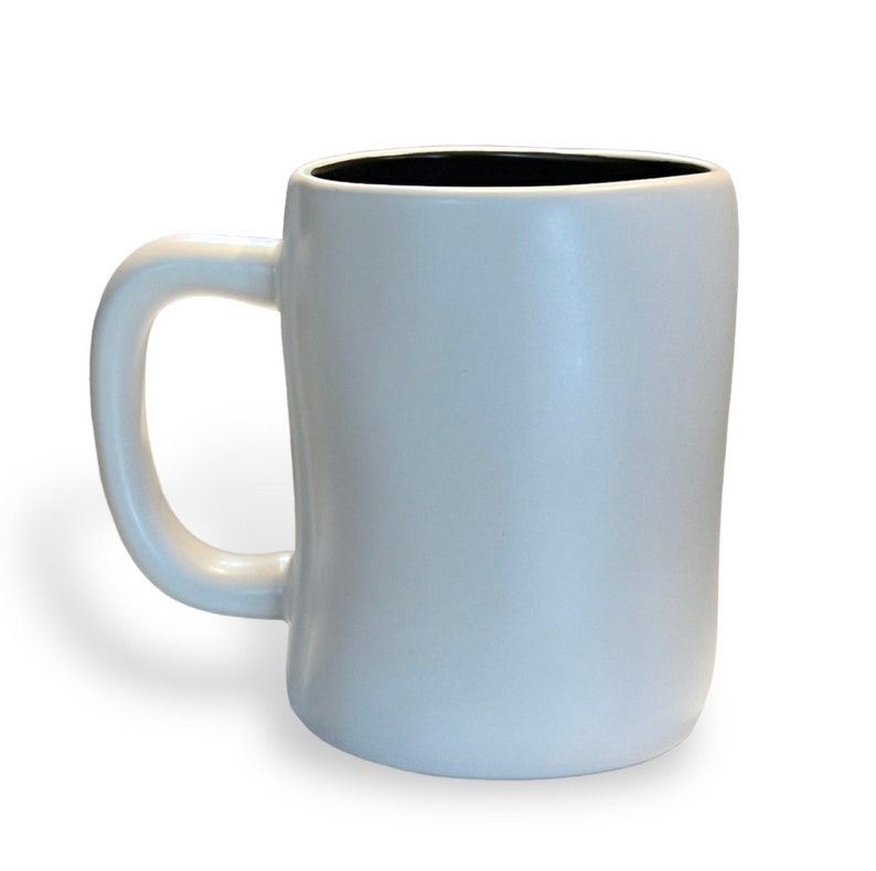 May include: A white ceramic mug with a black interior. The mug has a rounded rectangular shape with a curved handle. The mug is approximately 4 inches tall.