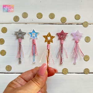 May include: Five tooth fairy wands with star-shaped tops and glitter. The wands are made of clear plastic and have a tooth design in the center of the star. Each wand has a pink tassel attached to the bottom. The wands are arranged in a row on a white wooden surface.