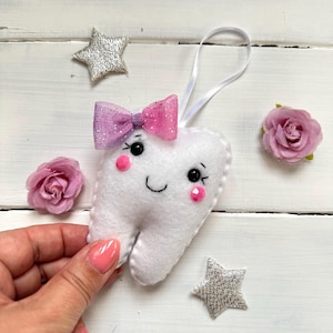 May include: A white felt tooth with a pink and purple bow, black eyes, and pink cheeks. The tooth is smiling and has a loop for hanging.