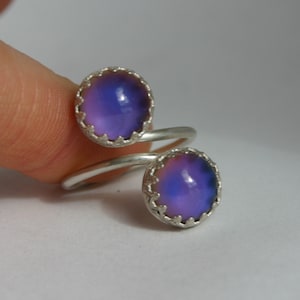May include: A silver-toned ring with two round, color-changing stones. The stones shift from purple to blue, set in ornate, scalloped bezels. The ring's band curves around the finger, creating a unique, eye-catching design.