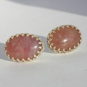 Può includere: Un paio di orecchini a bottone ovali. Ogni orecchino presenta una pietra preziosa rosa incastonata in una lunetta argentata smerlata. Le pietre preziose hanno un aspetto traslucido con sottili variazioni di colore.