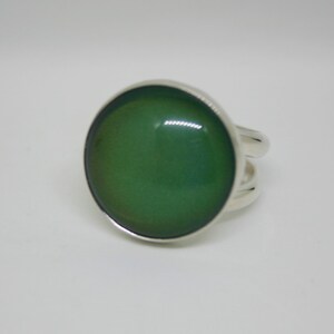 May include: A silver ring with a large, round, green mood stone. The ring has a simple band with a split design on each side of the stone, creating a modern look. The mood stone changes color.