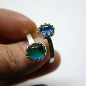 May include: Two silver rings with oval, color-changing stones. The stones shift from green to blue, set in a crown-like bezel. The rings are adjustable, with a simple band design. The rings are displayed against a white background.