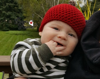 Puede incluir: Un bebé con un gorro rojo de ganchillo con una bellota de fieltro marrón en la parte superior. El bebé lleva una camisa a rayas y está siendo sostenido por alguien.