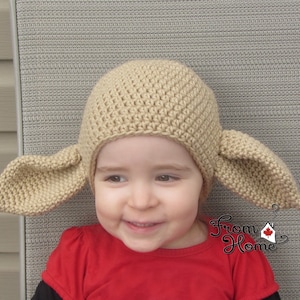 May include: A child wearing a beige crocheted hat with floppy ears. The hat is shaped like an elf's hat with pointed ears. The child is smiling and wearing a red shirt.