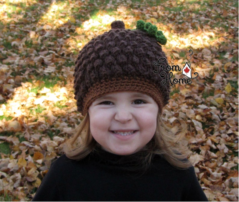 Puede incluir: Un ni&ntilde;o lleva un gorro de punto marr&oacute;n en forma de bellota con hojas verdes en la parte superior. El gorro est&aacute; hecho con un punto texturizado que se asemeja a la superficie abultada de una bellota. El ni&ntilde;o sonr&iacute;e y mira a la c&aacute;mara.