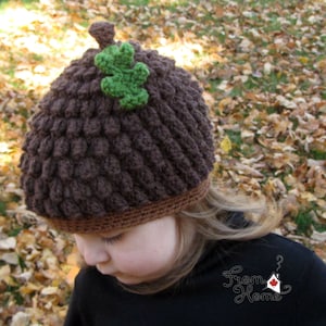 Puede incluir: Un gorro de punto marr&oacute;n en forma de bellota con una hoja verde en la parte superior. El gorro lo lleva puesto un ni&ntilde;o de pie en un mont&oacute;n de hojas de oto&ntilde;o.