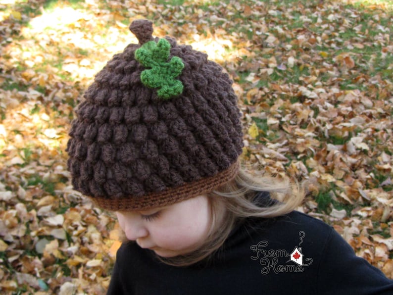 Puede incluir: Un gorro de punto marr&oacute;n en forma de bellota con una hoja verde en la parte superior. El gorro lo lleva puesto un ni&ntilde;o de pie en un mont&oacute;n de hojas de oto&ntilde;o.