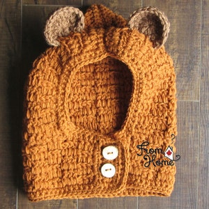 May include: A brown crocheted hooded garment with bear ears and two white buttons. The garment is made with a textured stitch and has a "From Home" logo on the right side.