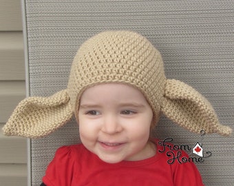Puede incluir: Un niño con un gorro de ganchillo beige con orejas caídas. El gorro tiene forma de gorro de elfo con orejas puntiagudas. El niño sonríe y lleva una camisa roja.
