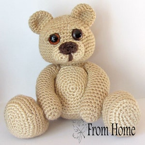 May include: A crocheted beige teddy bear with brown eyes and a brown nose. The bear is sitting with its legs crossed and its arms resting on its lap.