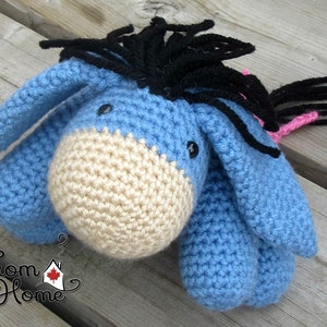 May include: A blue and beige crocheted donkey toy with black yarn for the mane and tail. The donkey has black eyes and a pink tail.