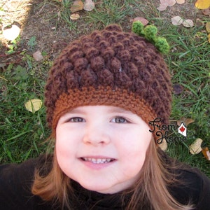 Puede incluir: Un ni&ntilde;o peque&ntilde;o con un gorro de ganchillo marr&oacute;n y naranja en forma de bellota con una hoja verde en la parte superior. El gorro tiene un dise&ntilde;o texturizado y abultado. El ni&ntilde;o sonr&iacute;e y mira a la c&aacute;mara.