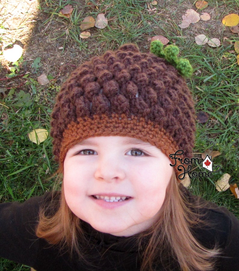 Puede incluir: Un ni&ntilde;o peque&ntilde;o con un gorro de ganchillo marr&oacute;n y naranja en forma de bellota con una hoja verde en la parte superior. El gorro tiene un dise&ntilde;o texturizado y abultado. El ni&ntilde;o sonr&iacute;e y mira a la c&aacute;mara.