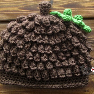 Puede incluir: Un gorro de punto de color marr&oacute;n en forma de bellota con una hoja de punto verde en la parte superior. El gorro est&aacute; hecho con un punto texturizado que se asemeja a las escamas de una bellota.