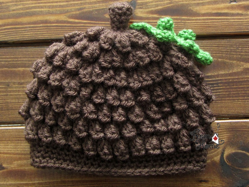 Puede incluir: Un gorro de punto de color marr&oacute;n en forma de bellota con una hoja de punto verde en la parte superior. El gorro est&aacute; hecho con un punto texturizado que se asemeja a las escamas de una bellota.