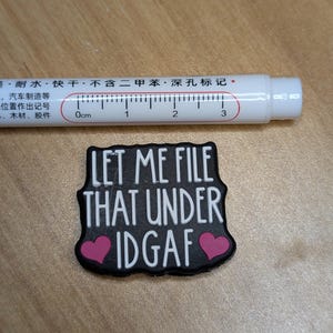 May include: A white pen with a ruler scale and text in Chinese characters is next to a black patch with the text "LET ME FILE THAT UNDER IDGAF" and two pink hearts.