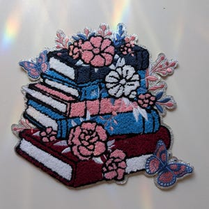 May include: Embroidered patch featuring a stack of books in shades of burgundy, blue, and navy, adorned with pink and white flowers and butterflies. The patch has a silver metallic border, creating a decorative and whimsical design.