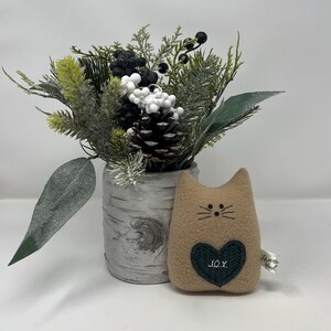 May include: A tan cat-shaped plush toy with embroidered whiskers and a green heart with the word "JOY". Beside it is a birch-style vase with faux greenery, berries, and a pinecone.