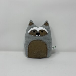 May include: Gray plush raccoon stuffed animal with black eyes, a white nose, and brown ears. The stuffed animal has a brown belly and a tag that says "Little Wood".