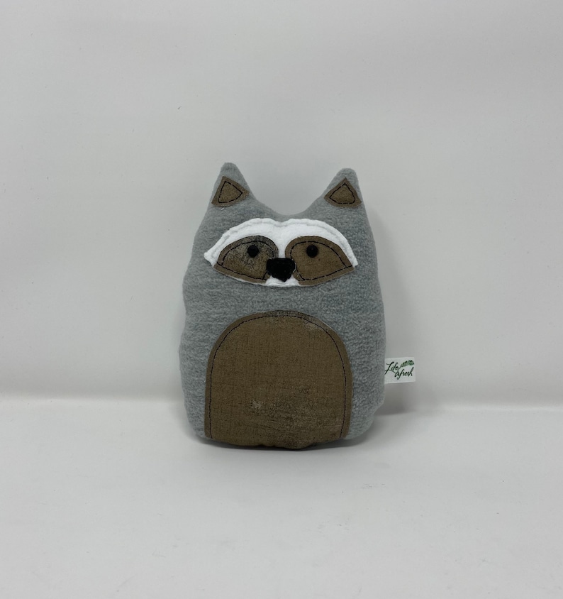 May include: Gray plush raccoon stuffed animal with black eyes, a white nose, and brown ears. The stuffed animal has a brown belly and a tag that says "Little Wood".