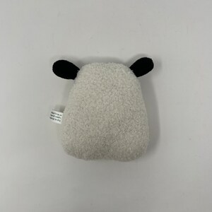 Pode incluir: Brinquedo de pel&uacute;cia branco em forma de ovelha com orelhas pretas. O brinquedo tem uma etiqueta que diz "Lavar na m&aacute;quina ou &agrave; m&atilde;o, pendurar para secar."