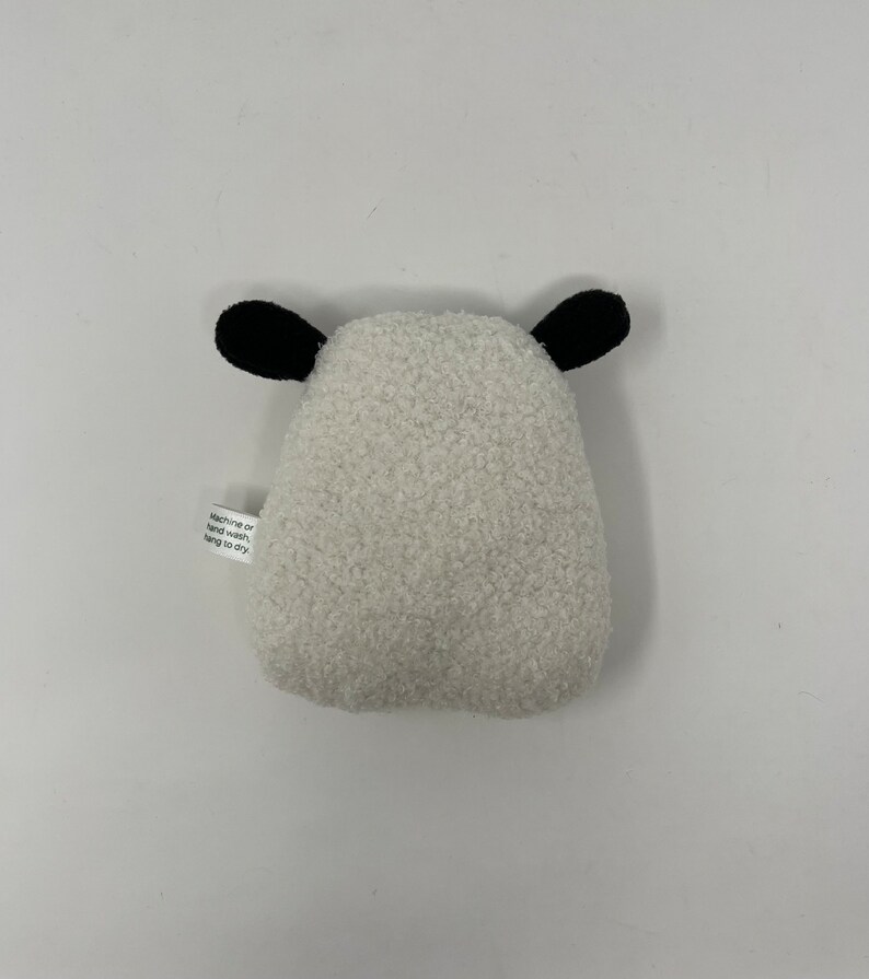 Pode incluir: Brinquedo de pel&uacute;cia branco em forma de ovelha com orelhas pretas. O brinquedo tem uma etiqueta que diz "Lavar na m&aacute;quina ou &agrave; m&atilde;o, pendurar para secar."