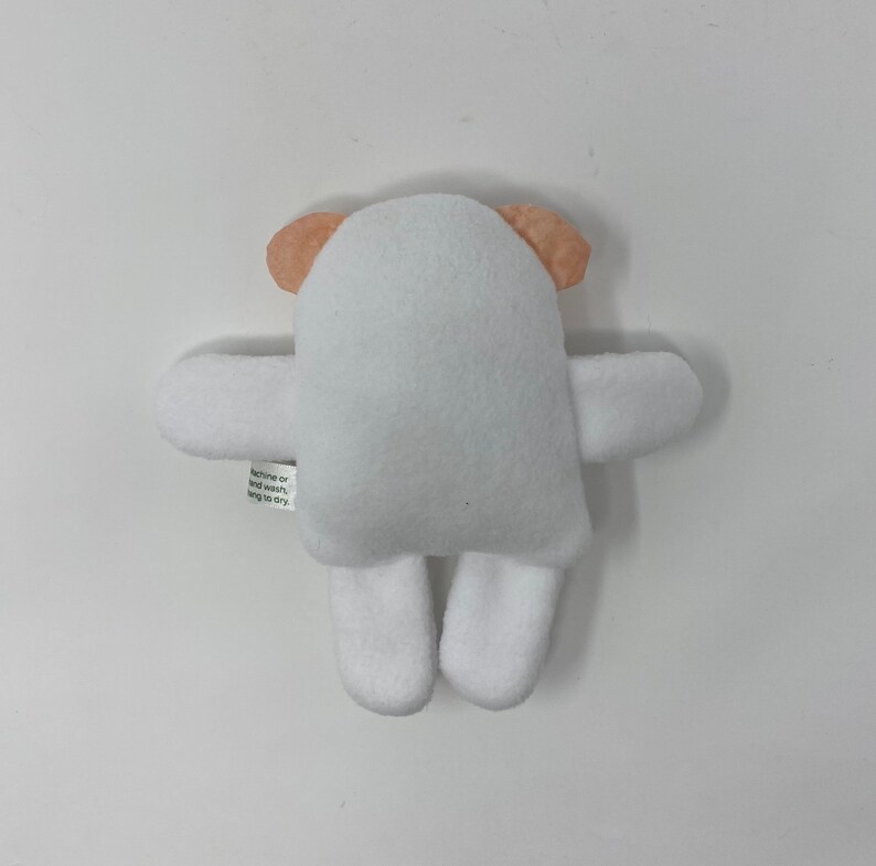 May include: White plush teddy bear with peach-colored ears. The bear has a simple design with no facial features. The text "machine or hand wash, hang to dry" is printed on the back of the bear.