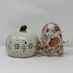 May include: A white ceramic pumpkin with a brown stem and a floral design. The pumpkin is next to a small, white ghost with a brown, orange and grey floral pattern. The ghost has the text "Little Spook" on the side.