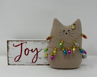 May include: A light brown plush cat figure decorated with a string of colourful Christmas lights. Beside it is a wooden sign with the word "Joy" in red lettering. The cat has embroidered facial features.