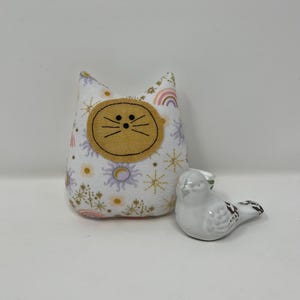 May include: A white fabric cat-shaped plush toy with a yellow cat face and a white ceramic bird with black spots. The cat toy has a pattern of rainbows, suns, and stars. The bird is small and has a smooth finish.