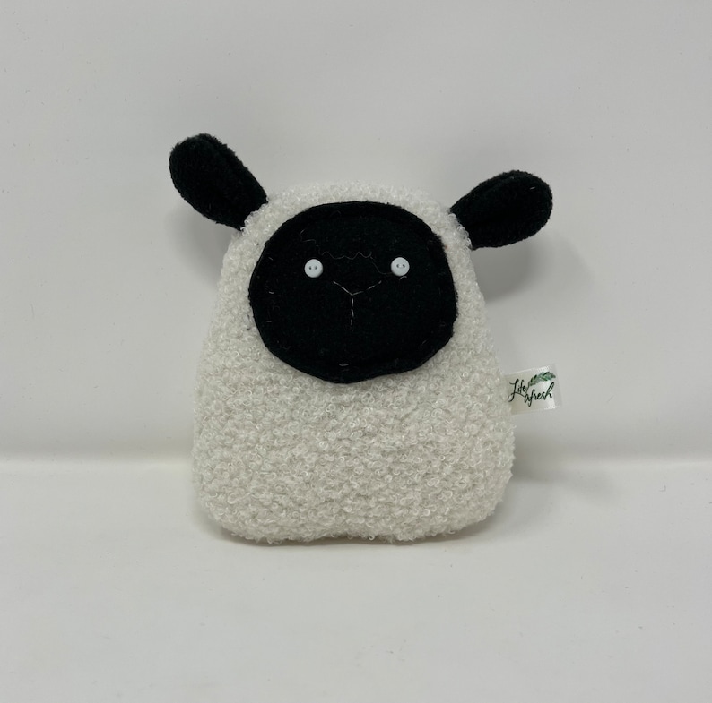 Pode incluir: Um brinquedo de pel&uacute;cia de ovelha branco com orelhas pretas, um rosto preto e olhos de bot&atilde;o brancos. O brinquedo tem uma pequena etiqueta verde com o texto "Little Fresh" nela.