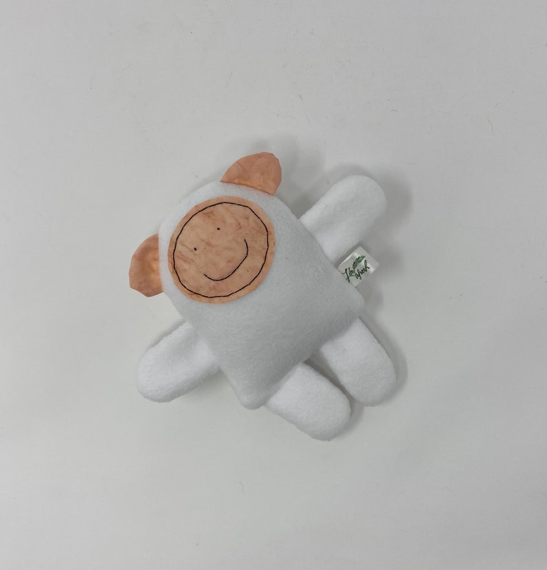 May include: White plush toy shaped like a person with a smiling face. The face is made of a brown fabric with a stitched smile. The toy has two small ears and two arms.