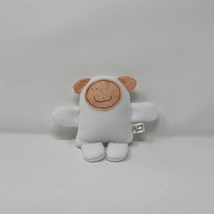 May include: White plush toy with a brown felt face and a smiling mouth. The toy has small ears and arms and is sitting on a white surface. The toy has a green tag with the text "Little Sprout" on it.