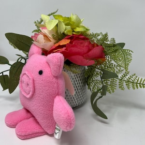 May include: A plush pink pig toy with a small tag, positioned in front of a vase of colorful flowers. The flowers include red, pink, and yellow blooms, with green foliage. The pig has small ears and a cute snout.