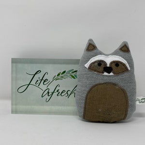 May include: Gray and brown plush raccoon toy with black eyes and a black nose. The toy is sitting on a white surface in front of a glass block with the words "life afresh" written on it.