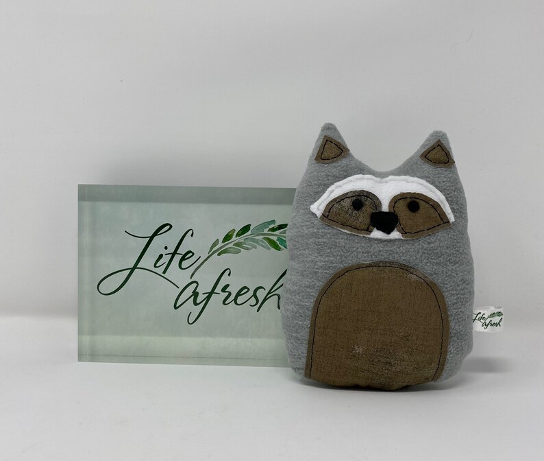 May include: Gray and brown plush raccoon toy with black eyes and a black nose. The toy is sitting on a white surface in front of a glass block with the words "life afresh" written on it.