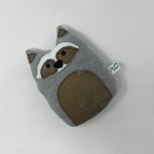 May include: A gray plush raccoon toy with black eyes, a black nose, and a brown belly. The toy has a white patch on its face and a small green tag that says "Little Bird".