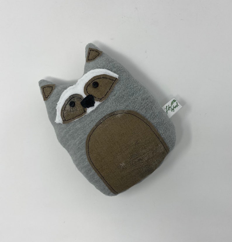 May include: A gray plush raccoon toy with black eyes, a black nose, and a brown belly. The toy has a white patch on its face and a small green tag that says "Little Bird".