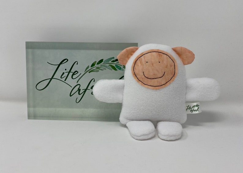 May include: White plush toy with a brown felt face and a smiling mouth. The toy has small ears and arms and is sitting on a white surface. The toy has a tag that says "Life/After/Life".