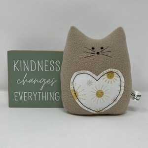 May include: A beige plush cat with a heart-shaped patch on its chest. The patch is white with a yellow sun pattern and the text "Kindness changes everything" is on a green wooden sign behind the cat.