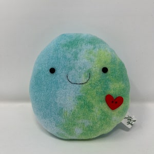 May include: A green and blue plush Earth with a smiling face and a red heart sewn on the side. The plush toy has a tag that says "Little" with a leaf design.