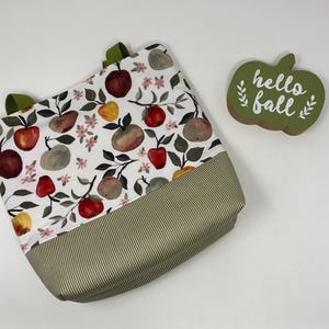 May include: A tote bag with a white background, featuring a colorful apple print and olive green handles. The bottom of the bag has a green and white striped pattern. A green wooden apple-shaped sign with the words "hello fall" is next to the bag.